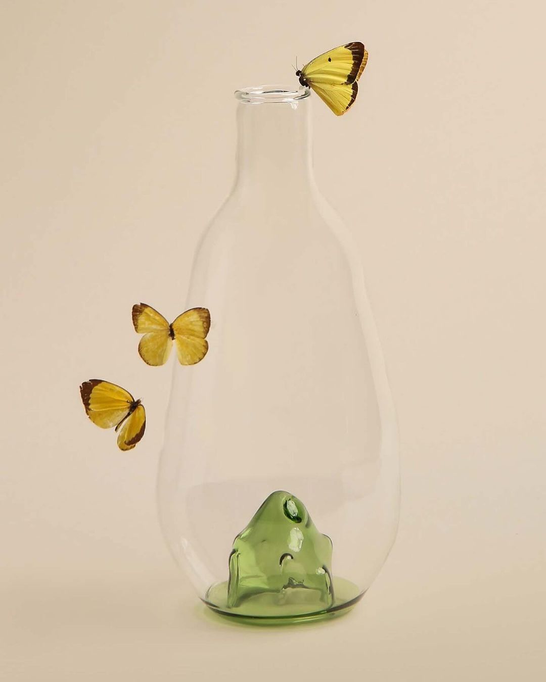 Still life of a clear glass vase with three yellow butterflies, one perched on top, against a cream-colored background in studio lighting.