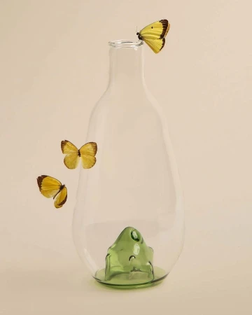Still life of a clear glass vase with three yellow butterflies, one perched on top, against a cream-colored background in studio lighting.
