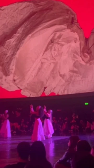 Performance art on stage with performers in flowing white costumes in front of a large seated audience and a screen above.