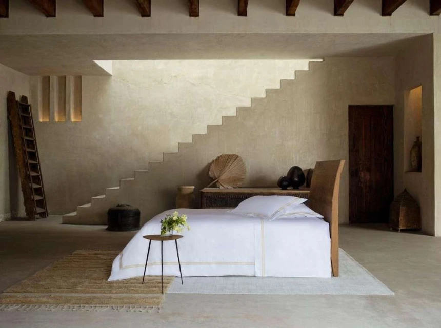 Interior of a bedroom with a bed, side table with flowers, neutral-toned walls, steps, wooden beams, and a ladder for a minimalist aesthetic.