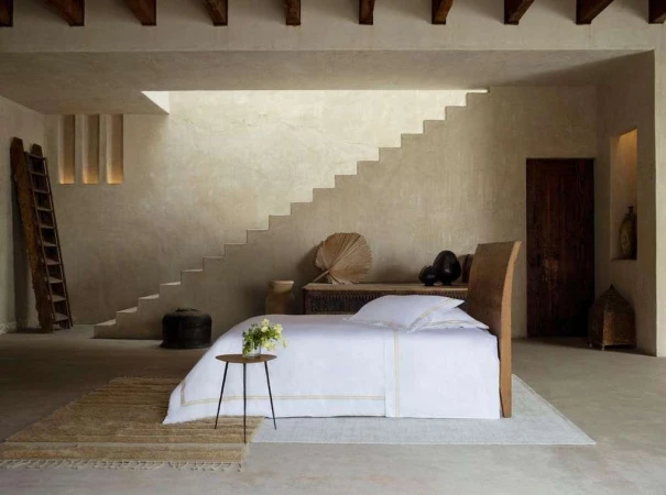 Interior of a bedroom with a bed, side table with flowers, neutral-toned walls, steps, wooden beams, and a ladder for a minimalist aesthetic.