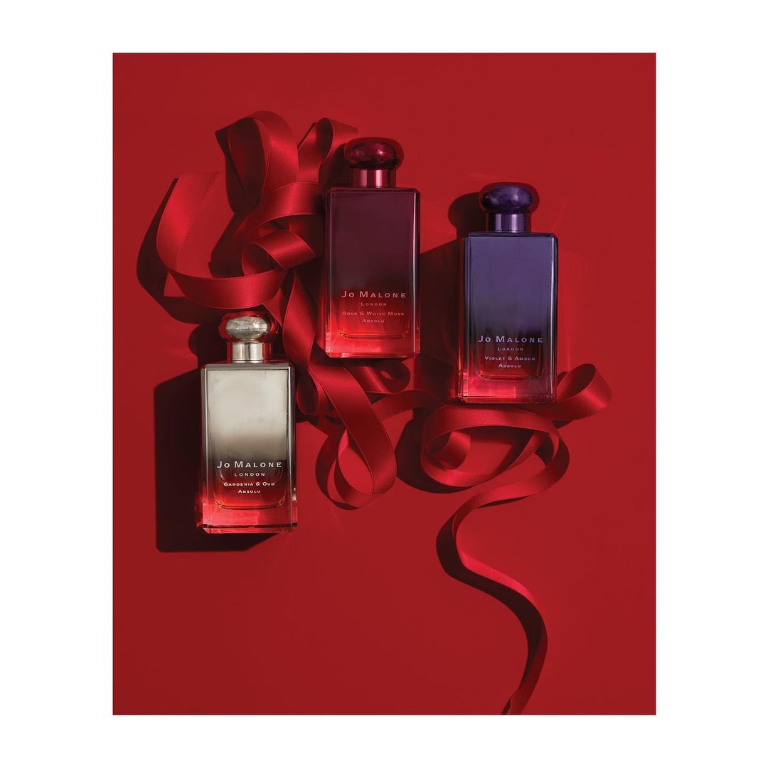 Three Jo Malone perfume bottles in red, purple, and silver sit on a red background with a red ribbon creating an elegant, festive arrangement.