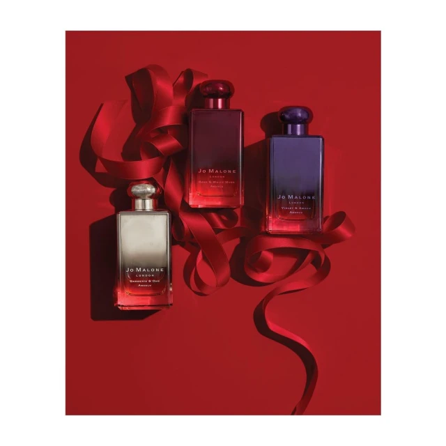 Three Jo Malone perfume bottles in red, purple, and silver sit on a red background with a red ribbon creating an elegant, festive arrangement.