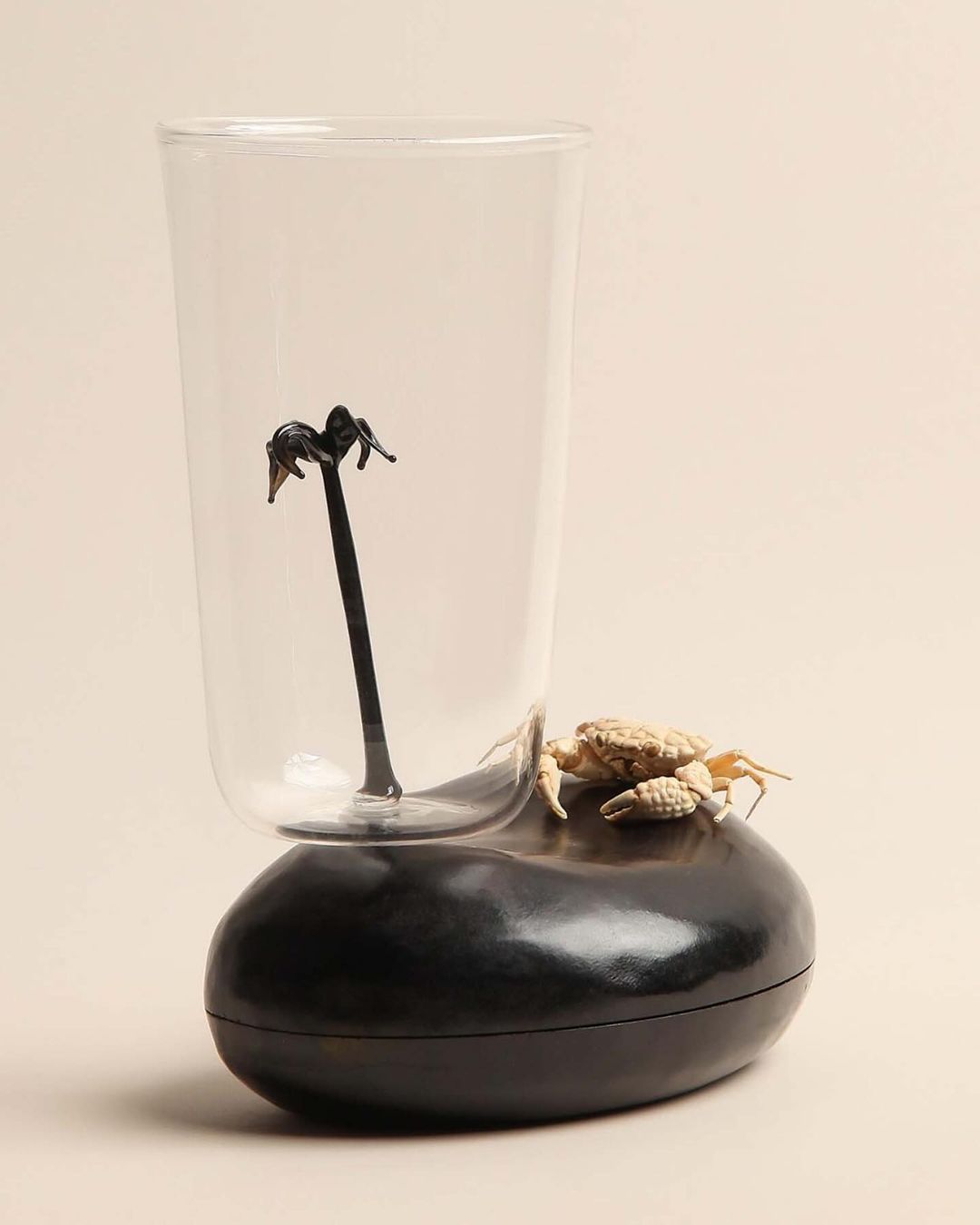 Still life of a glass with a stirrer, a small crab figurine, and a black, textured platform against a cream-colored backdrop.