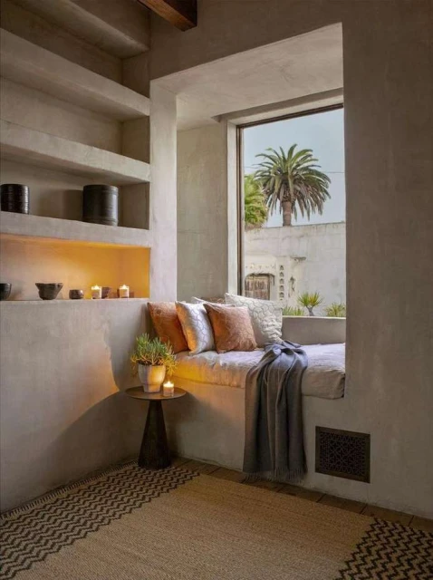 Cozy window seat nook with cushions and a blanket, beside a small round side table with a potted plant and candles, in a neutral-toned room.