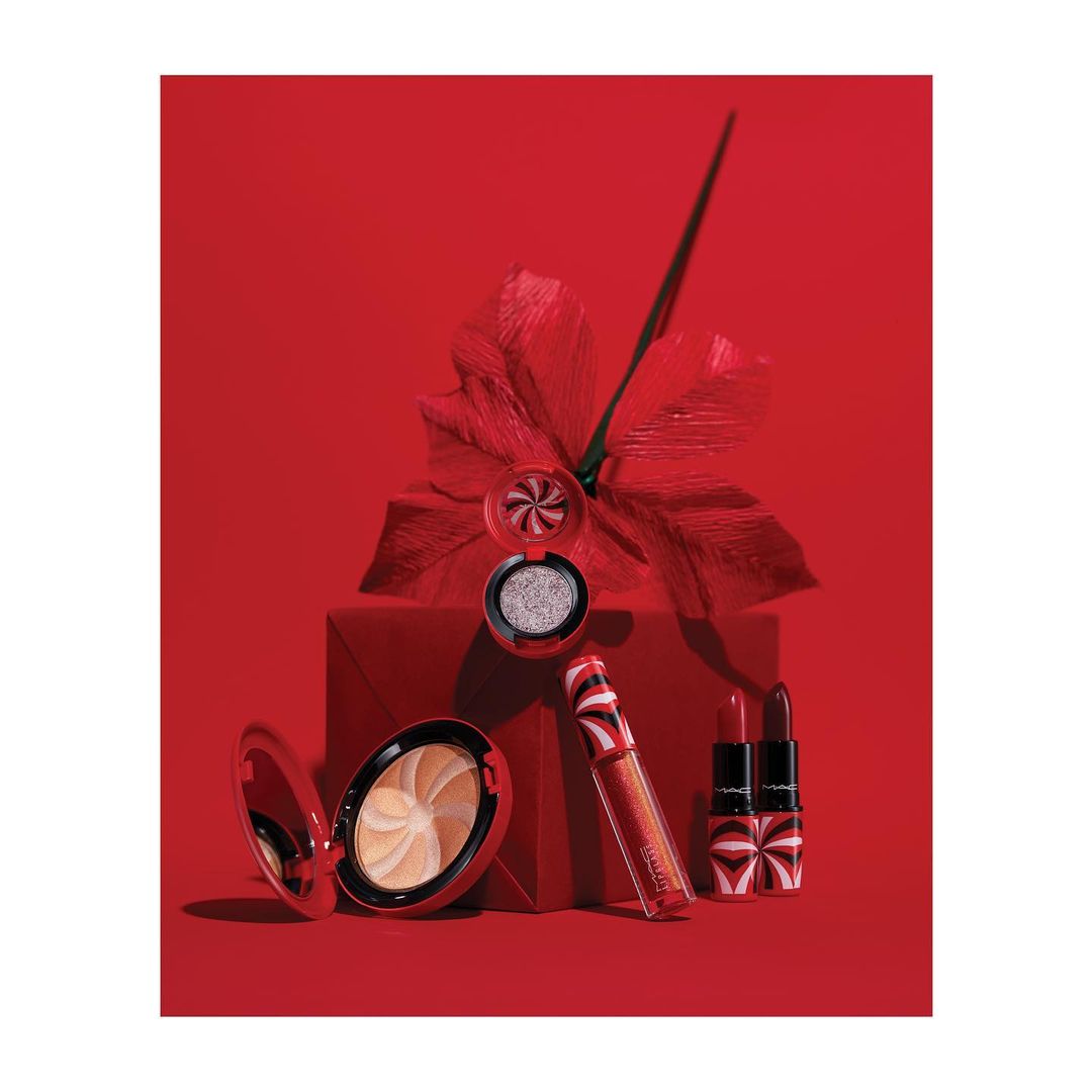 A collection of MAC cosmetics including lipstick, lip gloss, eyeshadow, and a powder compact arranged on and around a red gift box.