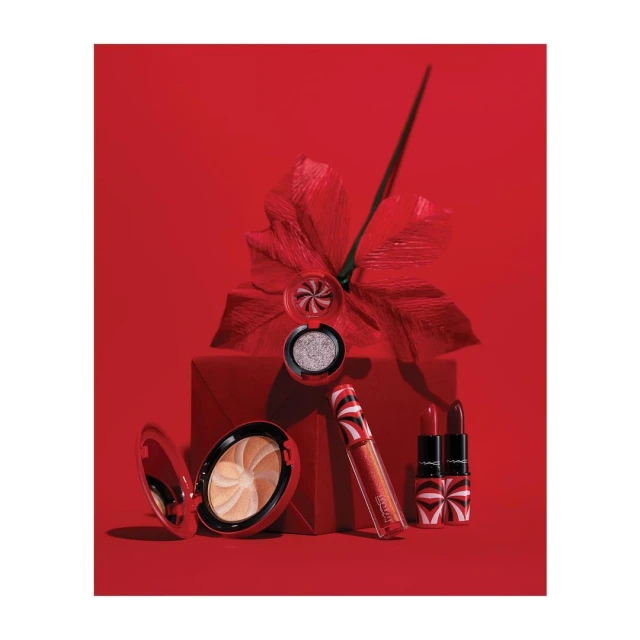 A collection of MAC cosmetics including lipstick, lip gloss, eyeshadow, and a powder compact arranged on and around a red gift box.