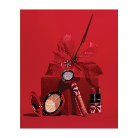 A collection of MAC cosmetics including lipstick, lip gloss, eyeshadow, and a powder compact arranged on and around a red gift box.