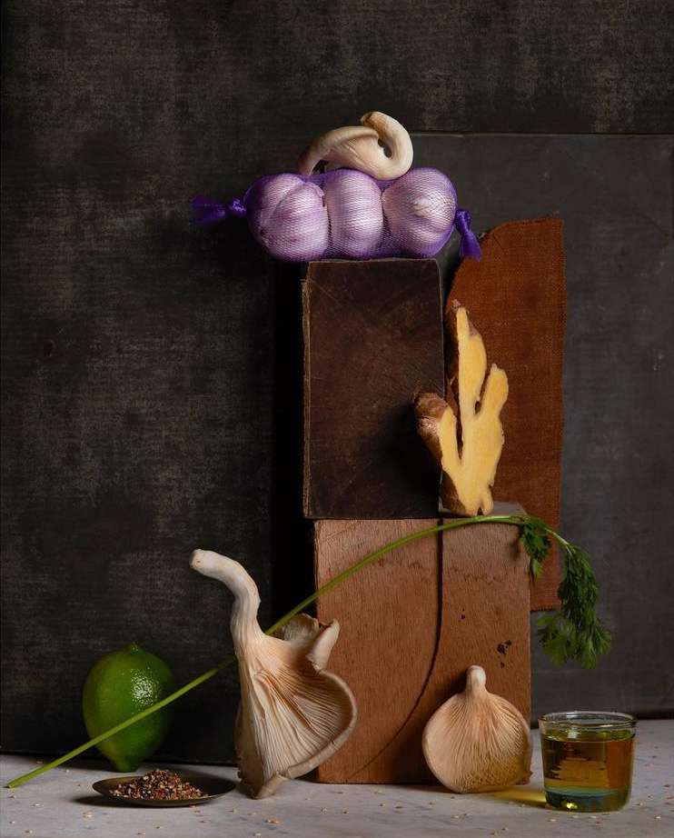 Still life featuring oyster mushrooms, garlic, lime, ginger, and olive oil arranged on wooden blocks against a dark background.