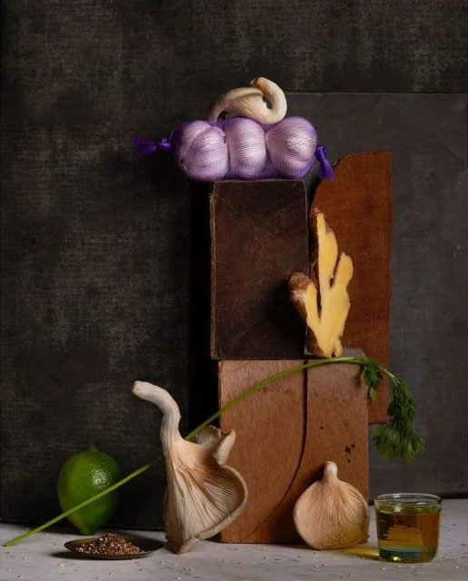 Still life featuring oyster mushrooms, garlic, lime, ginger, and olive oil arranged on wooden blocks against a dark background.