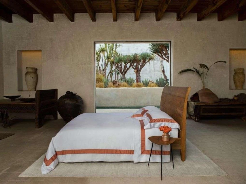 Bedroom interior featuring a bed with white linens and terracotta trim, side table with flowers, and a window view of desert plants.