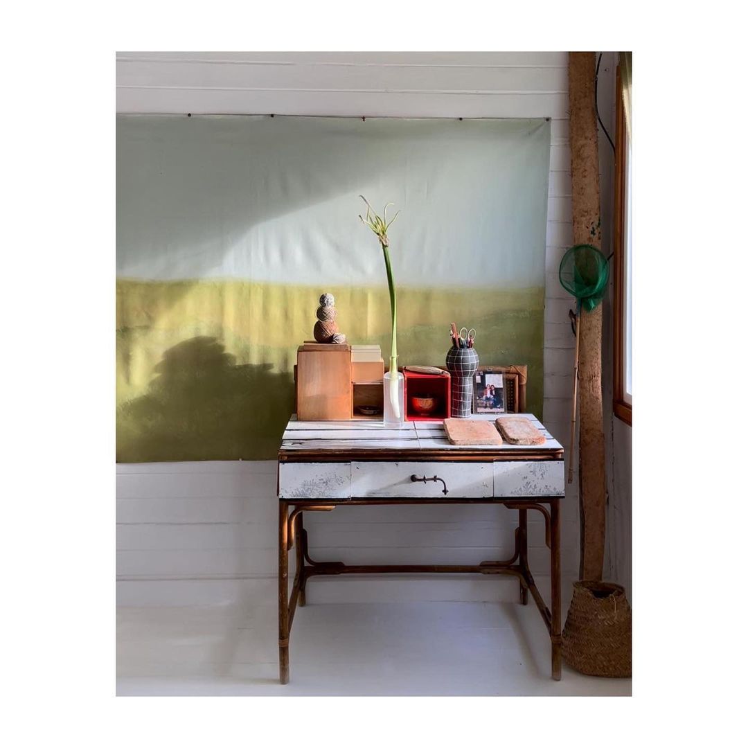 Indoor studio setup featuring a rustic desk with wooden boxes, vases, and a backdrop with a landscape-style painting in soft, muted tones.