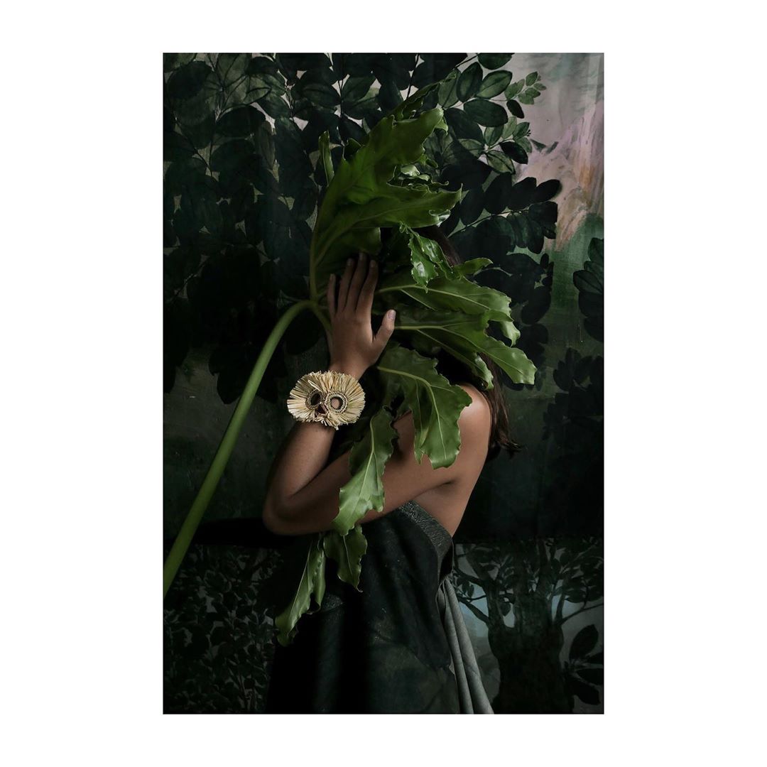 Woman with decorative golden bracelet poses partially hidden behind a large leaf in front of a green, leafy backdrop.