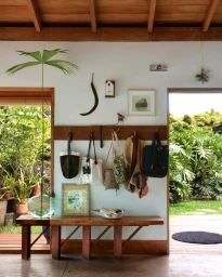Wooden hallway bench beneath a wall rack holding multiple bags and framed artworks in an airy interior with open doorways to lush greenery