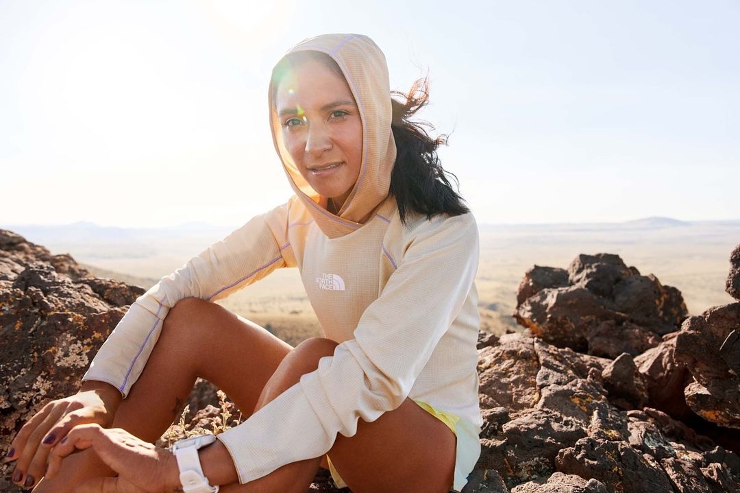 Female athlete in hooded shirt sits on rocky summit overlooking desert terrain, wearing watch and athletic gear under bright daylight sun.