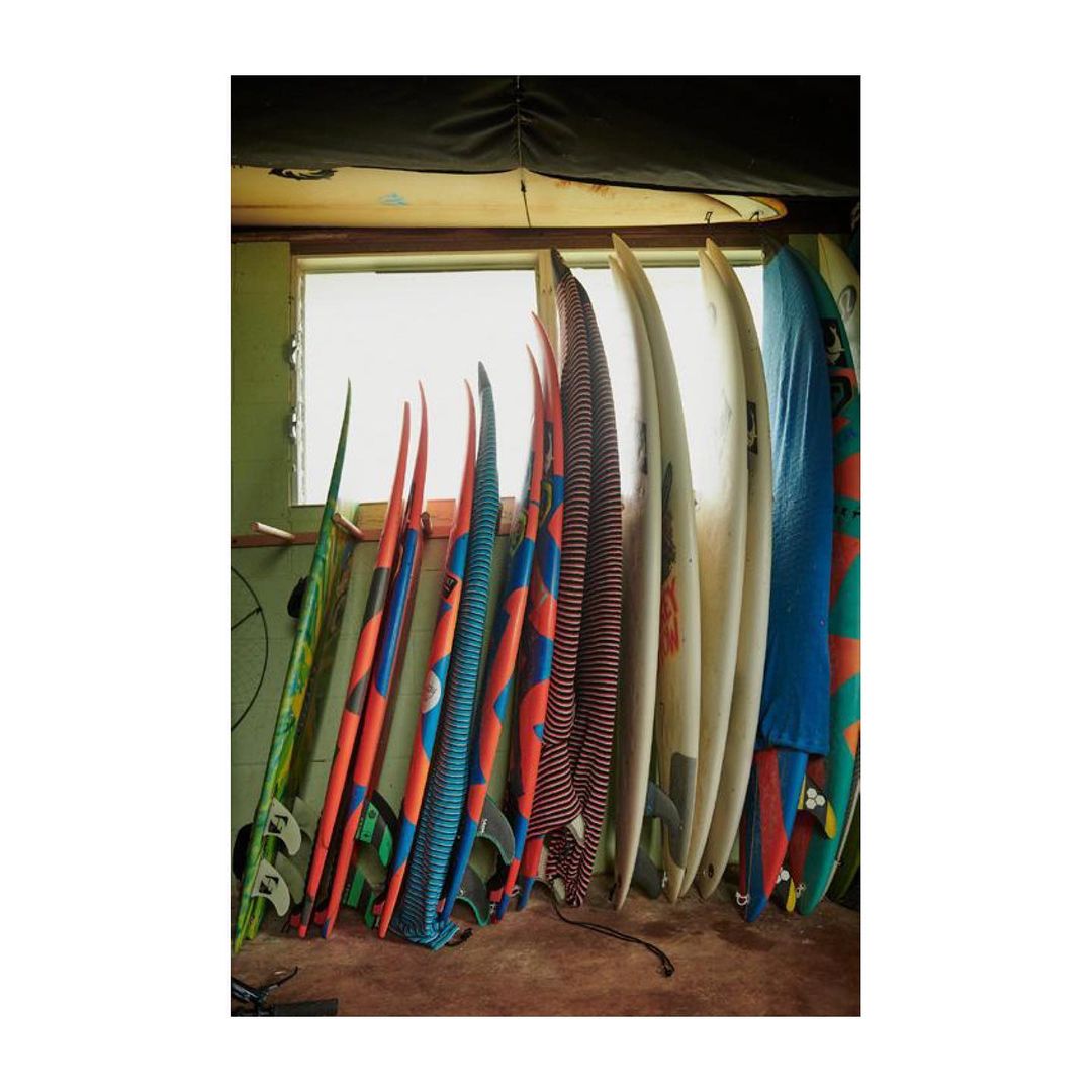 Array of colorful surfboards leaning against a wall inside a building, casting shadows on the ground; window behind them.