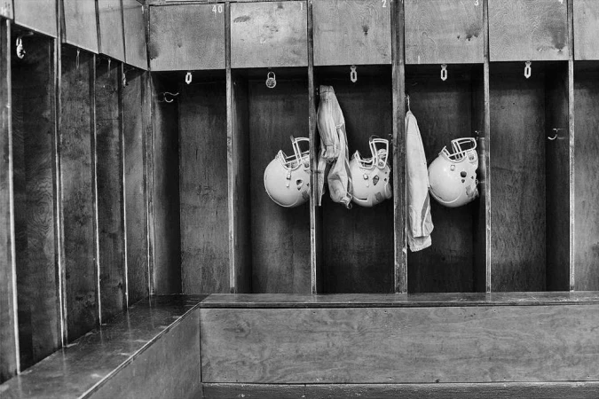 Black and white photograph of football helmets hanging in lockers, evoking a vintage or historical sports atmosphere.