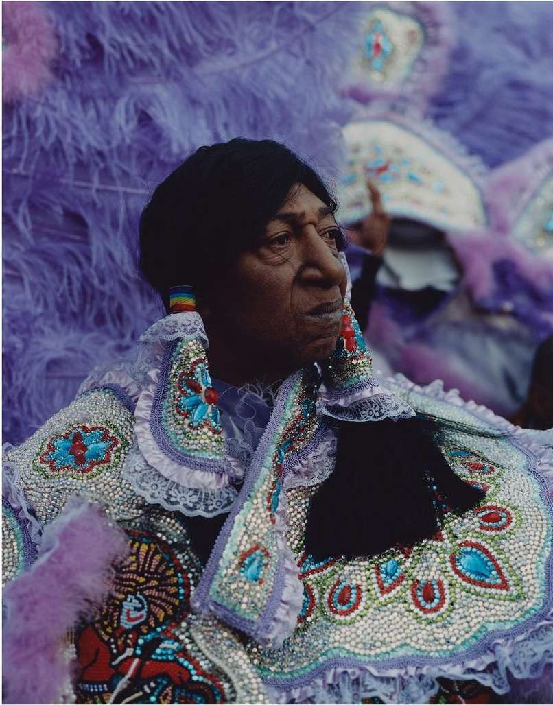 Portrait of an older woman in an embellished costume with feathers, beads, and lace, and a feather boa in shades of purple.