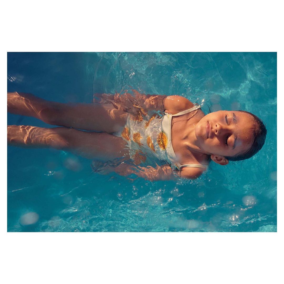 Young girl wearing a swimsuit floats on her back in a swimming pool with blue water, sunlight creating light and shadow patterns.