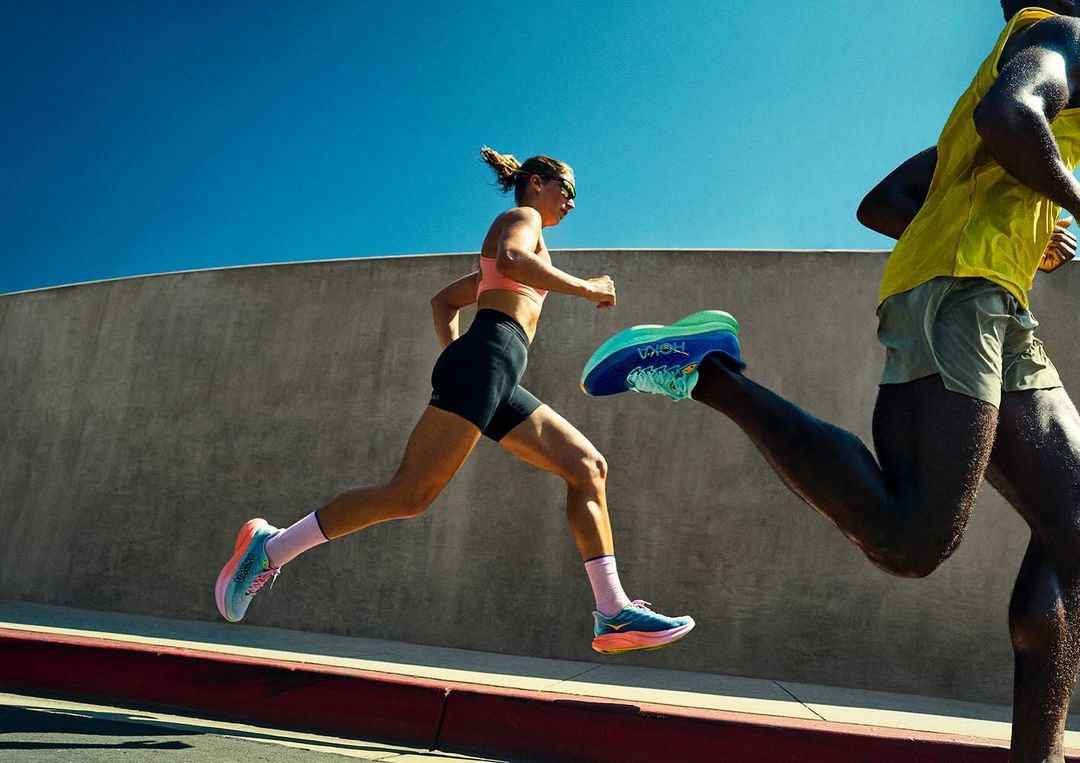Two runners sprint on the road wearing athletic apparel and Hoka brand running shoes under a clear sky with a concrete wall backdrop.