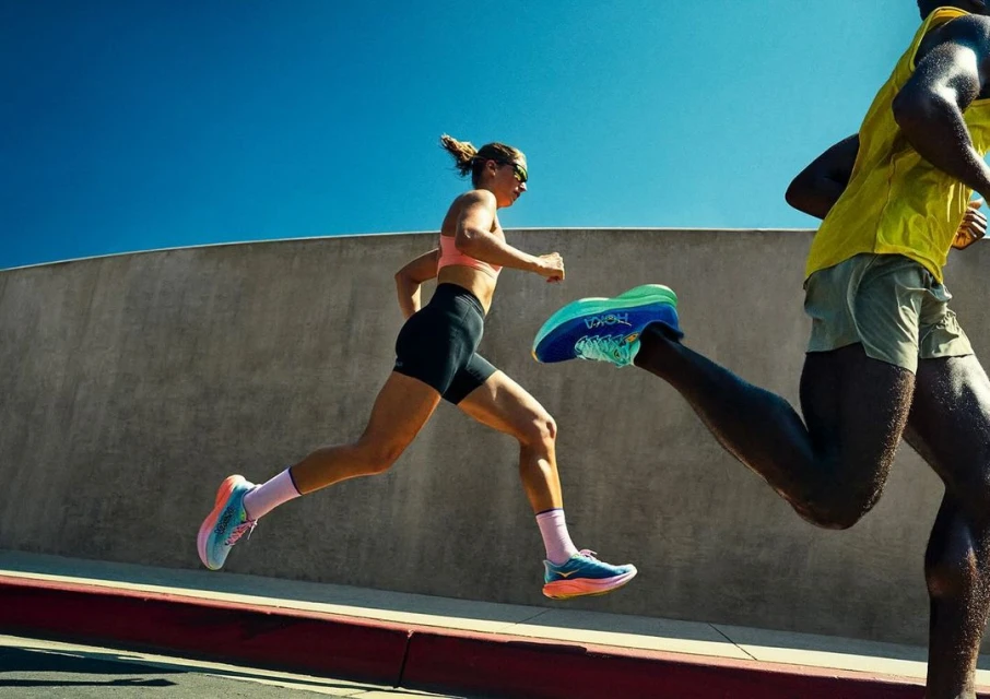 Two runners sprint on the road wearing athletic apparel and Hoka brand running shoes under a clear sky with a concrete wall backdrop.