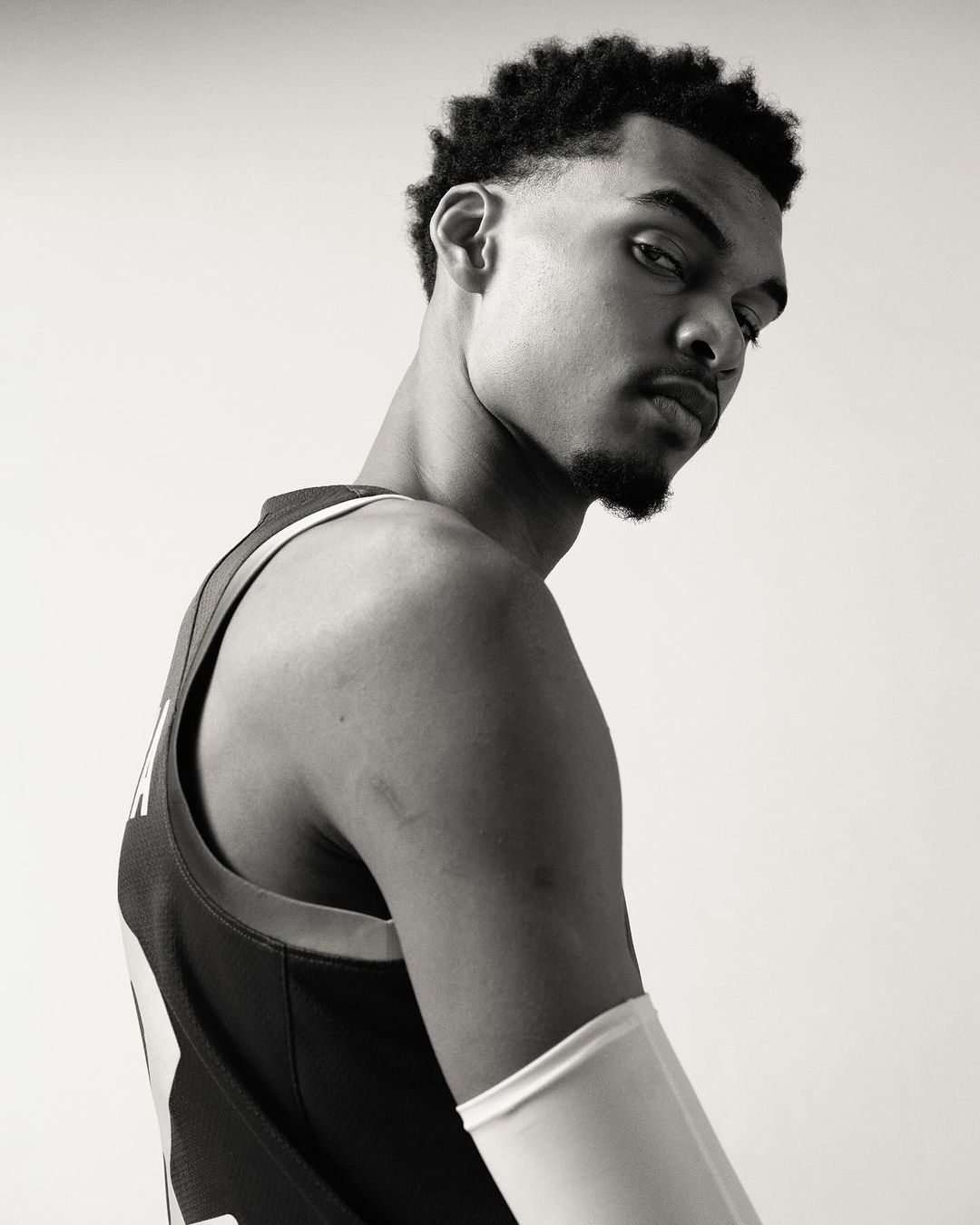 Black and white portrait of a young adult male athlete in a jersey, looking over his shoulder in a photo studio setting.
