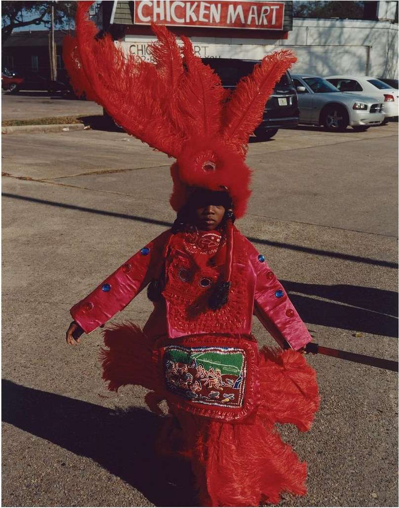 Child in elaborate red feathered costume stands on a street in front of a Chicken Mart, creating a vibrant and captivating scene.