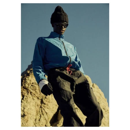 Man wearing a blue jacket, black beanie, and sunglasses standing outdoors against a rocky terrain under a clear sky.