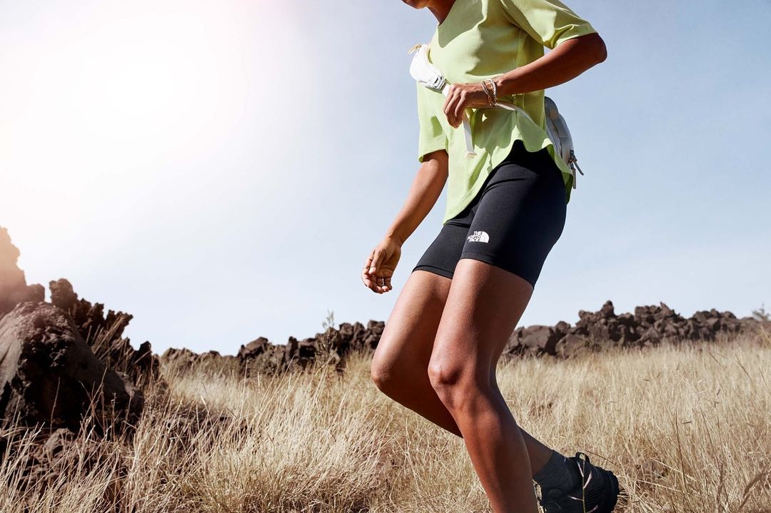 Female athlete in athletic shorts and a neon green T-shirt trail running on a grassy trail with rocky terrain in the background on a sunny day.