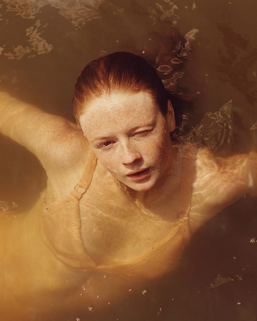 A woman with freckles and red hair stands in brown water, wearing a tan top. She gazes directly at the viewer from a high-angle perspective.
