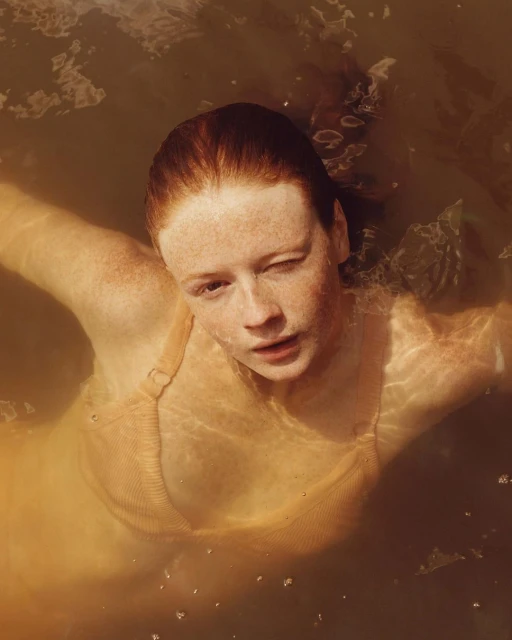A woman with freckles and red hair stands in brown water, wearing a tan top. She gazes directly at the viewer from a high-angle perspective.