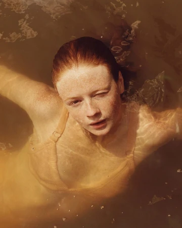 A woman with freckles and red hair stands in brown water, wearing a tan top. She gazes directly at the viewer from a high-angle perspective.