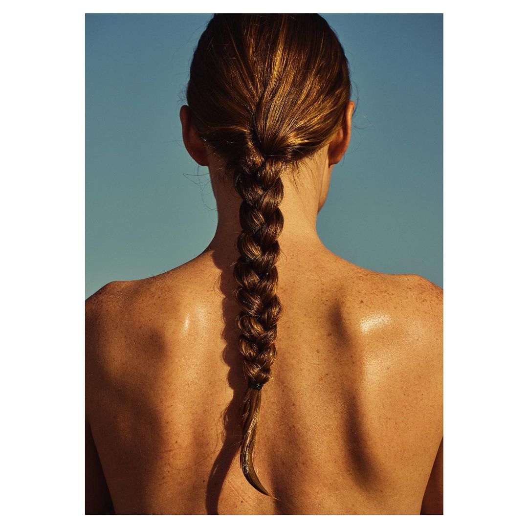Rear view of a person with a long braided hairstyle and sunlit bare shoulders against a clear blue sky background.