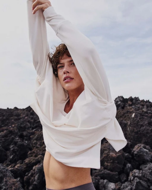 A young adult woman in a sweatshirt stretches her arms overhead outdoors against a rocky, volcanic background and cloudy sky.