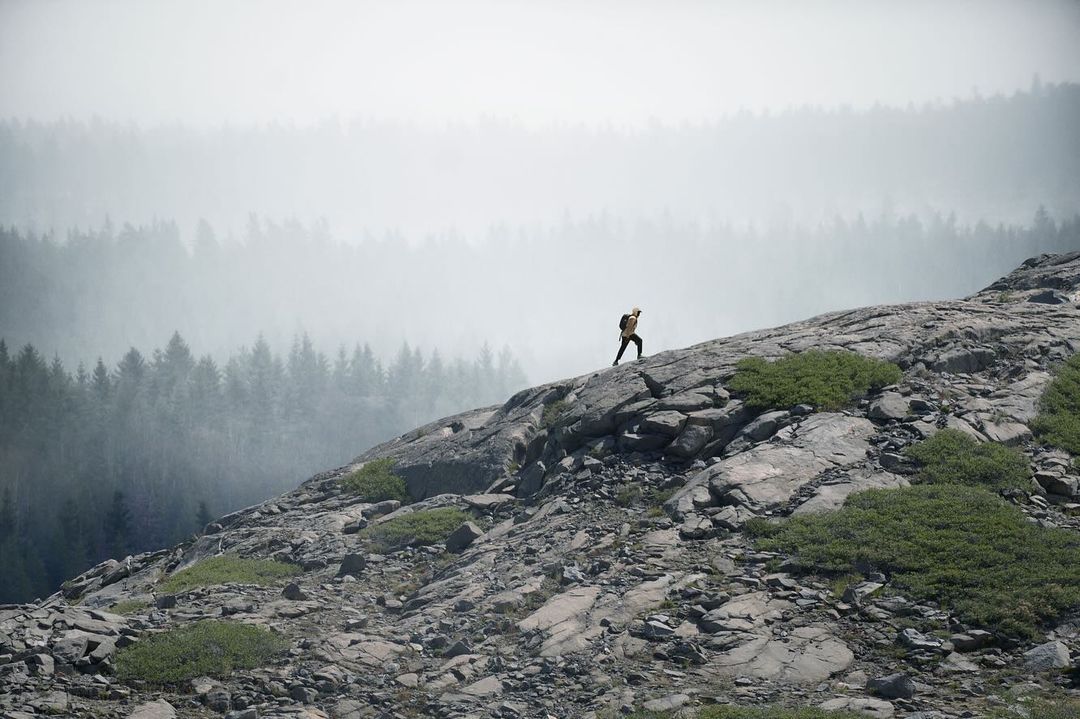 A hiker climbs a rocky slope in a mountainous landscape, with trees visible through the fog in the background. The scene is natural and serene.