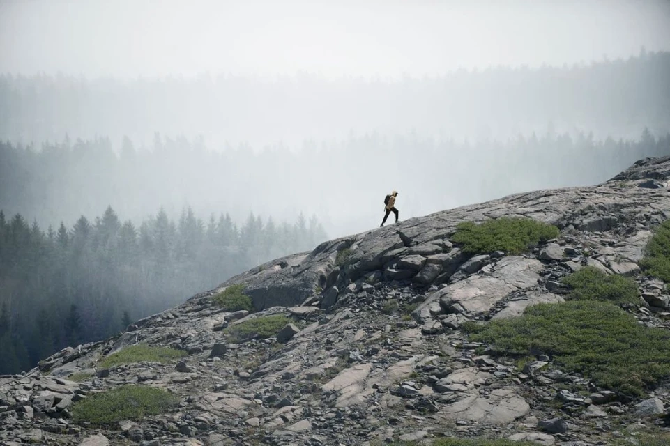A hiker climbs a rocky slope in a mountainous landscape, with trees visible through the fog in the background. The scene is natural and serene.
