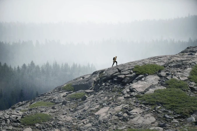 A hiker climbs a rocky slope in a mountainous landscape, with trees visible through the fog in the background. The scene is natural and serene.