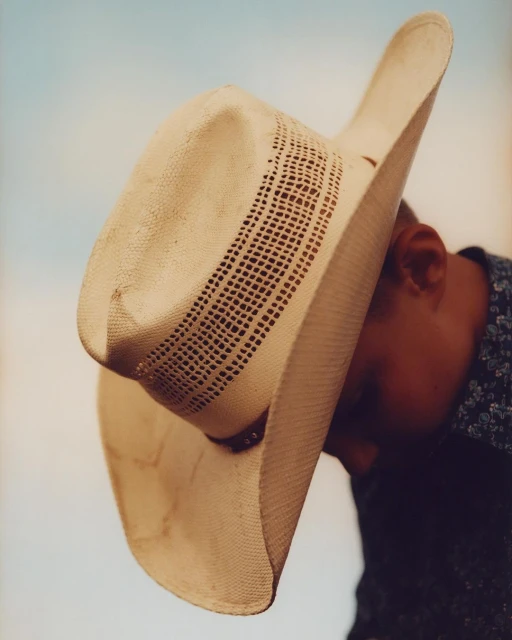 Close-up of a tan cowboy hat tilted over a person's face, obscuring the features, with the person wearing a patterned shirt.