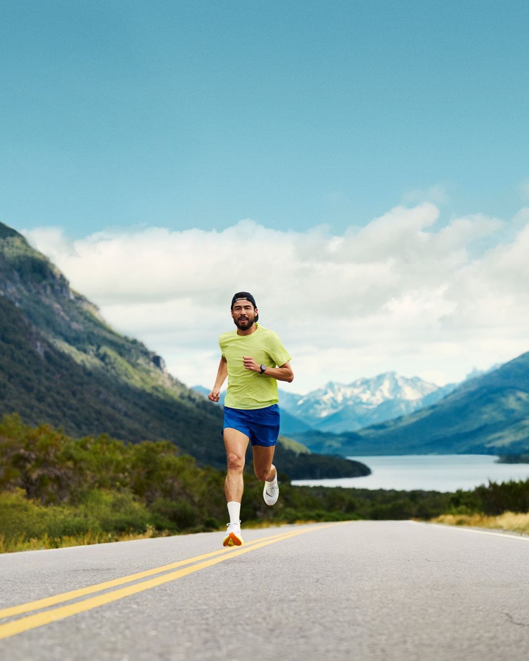 Male runner in athletic wear runs on rural asphalt road with mountains and blue sky in the background, enhancing travel or fitness concept.