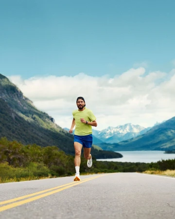 Male runner in athletic wear runs on rural asphalt road with mountains and blue sky in the background, enhancing travel or fitness concept.
