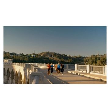 Group of women in athletic wear jogging across a concrete bridge structure on a sunny day, with a backdrop of green hills.