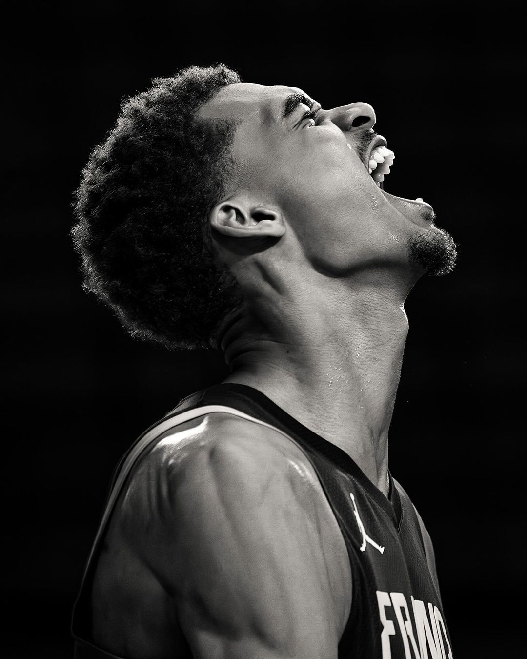Monochrome portrait of an African-American male athlete in a France jersey, shouting with an open mouth, head tilted back against a dark backdrop.