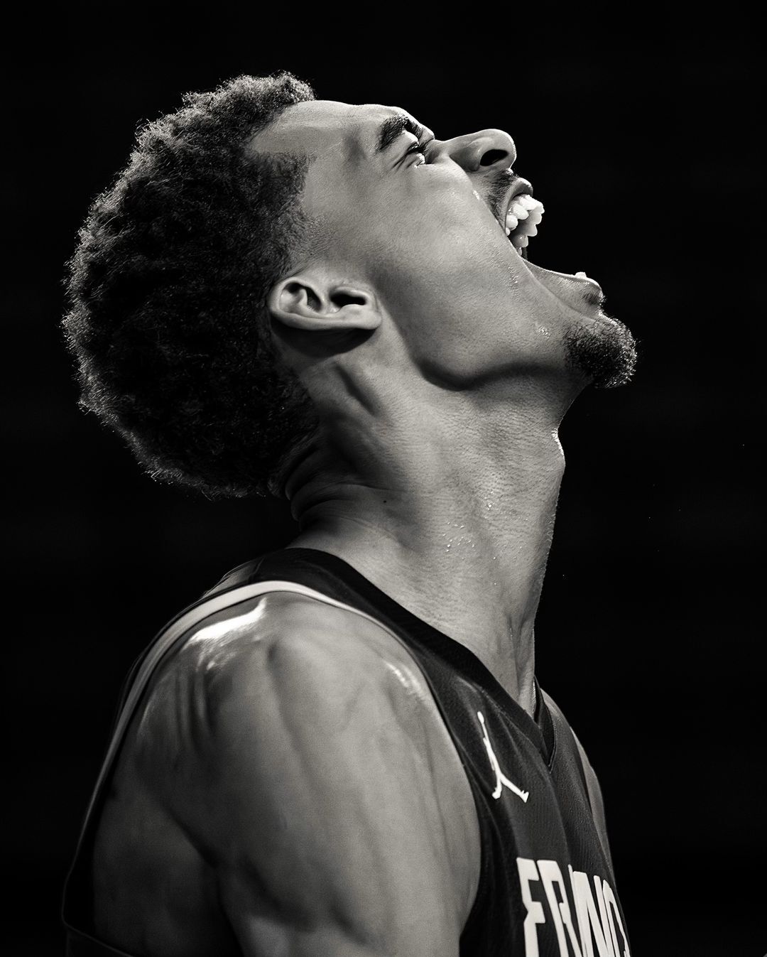 Black and white portrait of a male basketball player with his mouth open, wearing a France jersey with a dark background.
