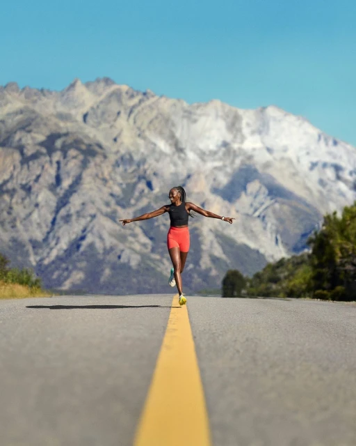 Athletic woman balances on a yellow line on a road with mountains in the background. Sunny day and blue sky.