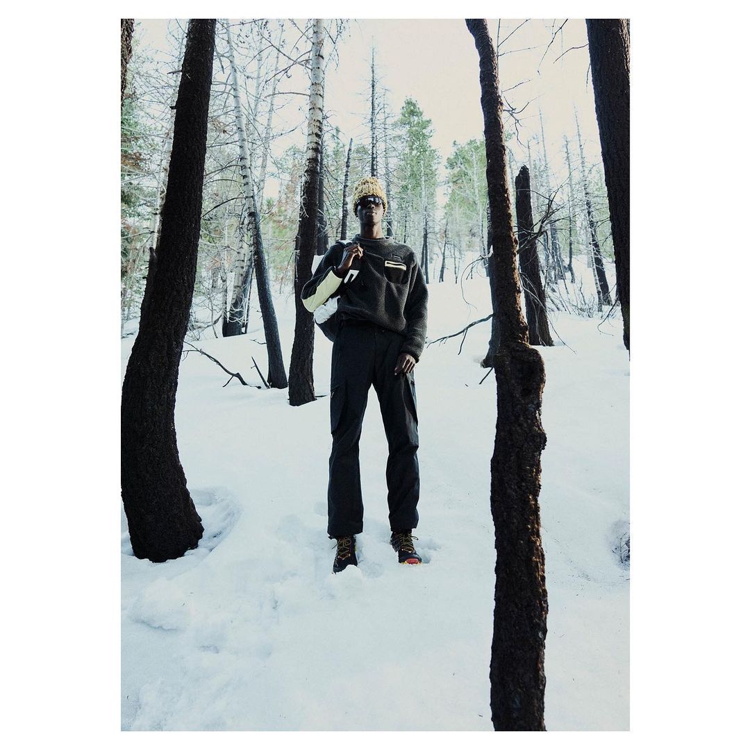 Adult male in a dark pullover and pants stands in a snowy forest with bare trees and tree trunks in the foreground, with a white backpack.