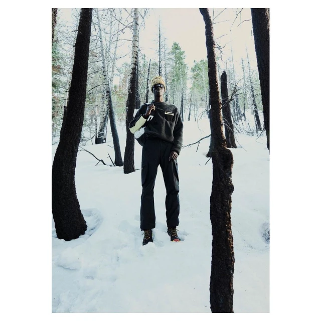 Adult male in a dark pullover and pants stands in a snowy forest with bare trees and tree trunks in the foreground, with a white backpack.