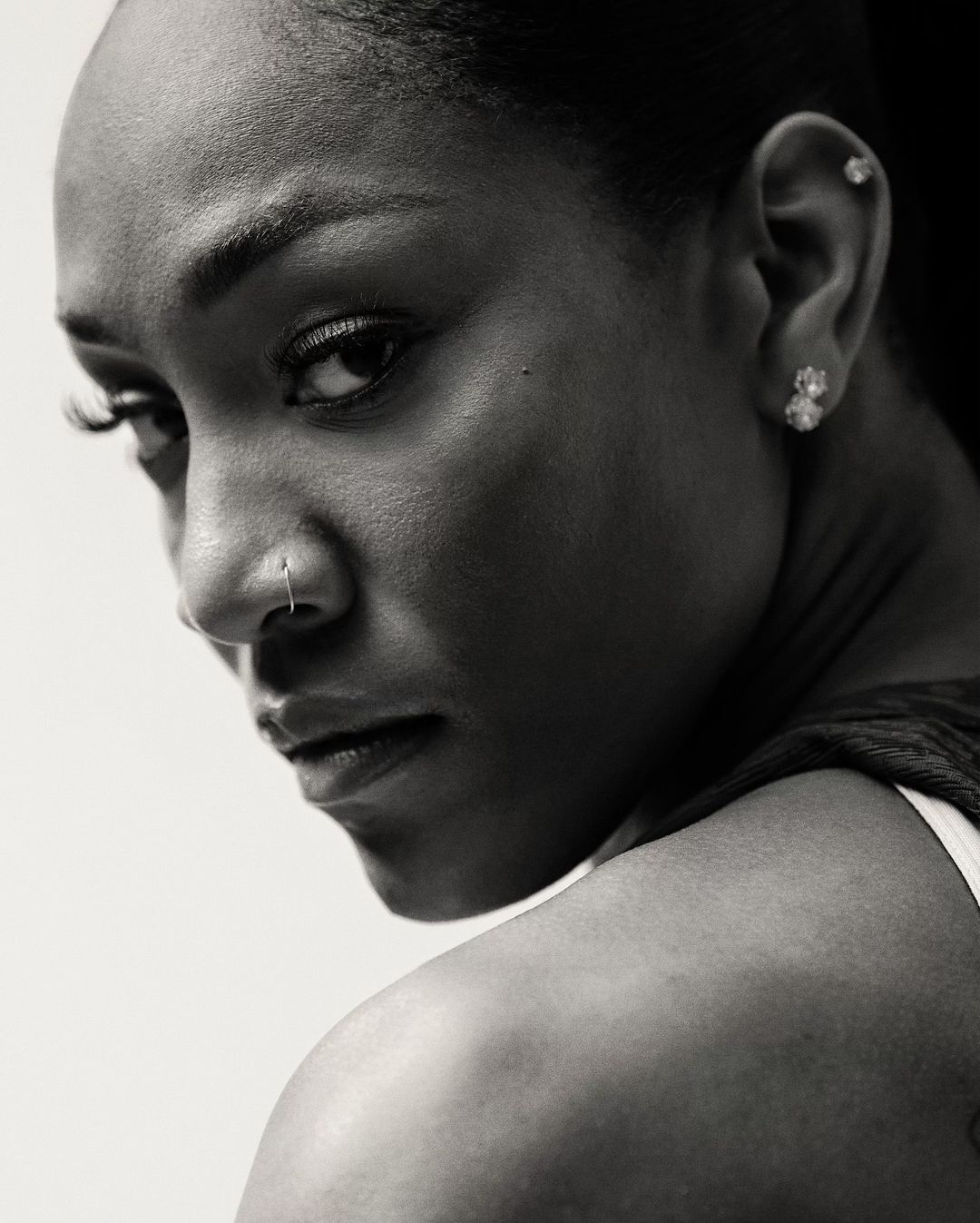 Close-up studio portrait of a woman with dark hair glancing over her shoulder; she has a nose ring and earrings with a neutral expression.