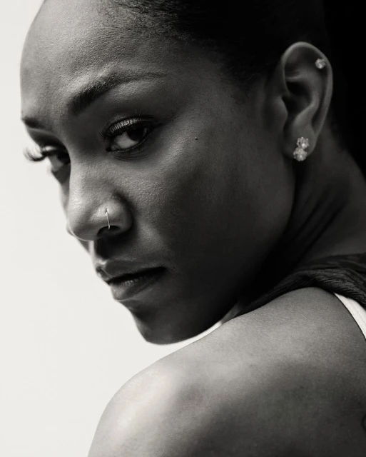 Close-up studio portrait of a woman with dark hair glancing over her shoulder; she has a nose ring and earrings with a neutral expression.