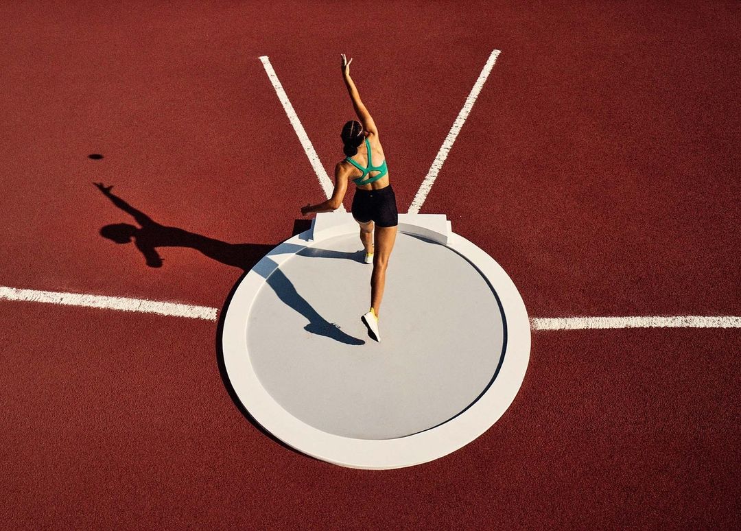 Overhead shot of female athlete in green sports bra about to throw shot put on red track with white lines on a sunny day.