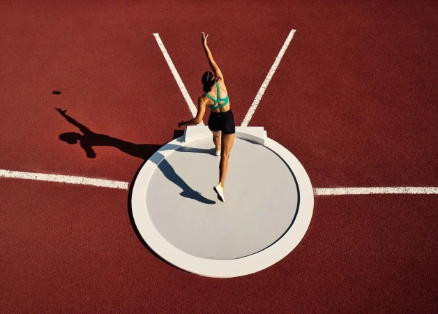 Overhead shot of female athlete in green sports bra about to throw shot put on red track with white lines on a sunny day.
